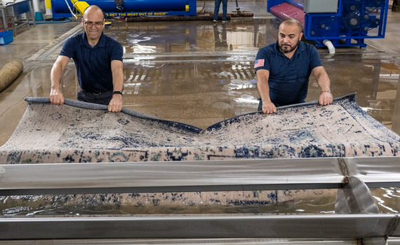 Two men professionally cleaning a rug using specialized equipment in an industrial setting.